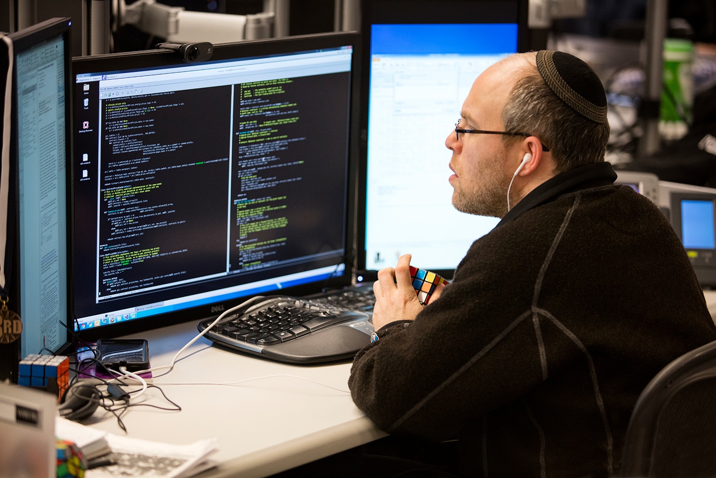 Man wearing kippah and headphones looks at code at his workstation while playing with Rubik’s Cubes.
