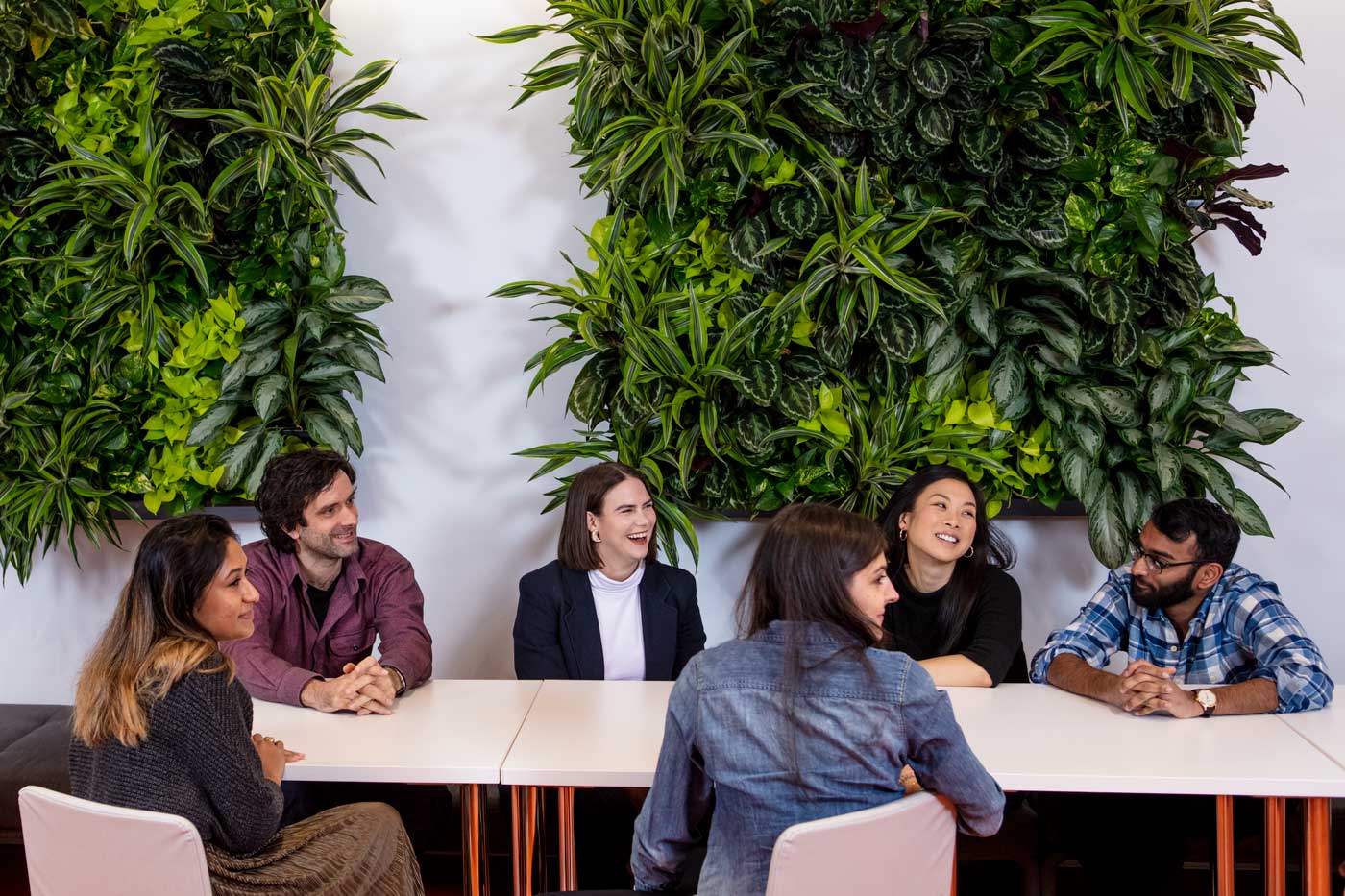 Six colleagues sit together around a table, talking and laughing in front of a plant wall in one of the town squares.